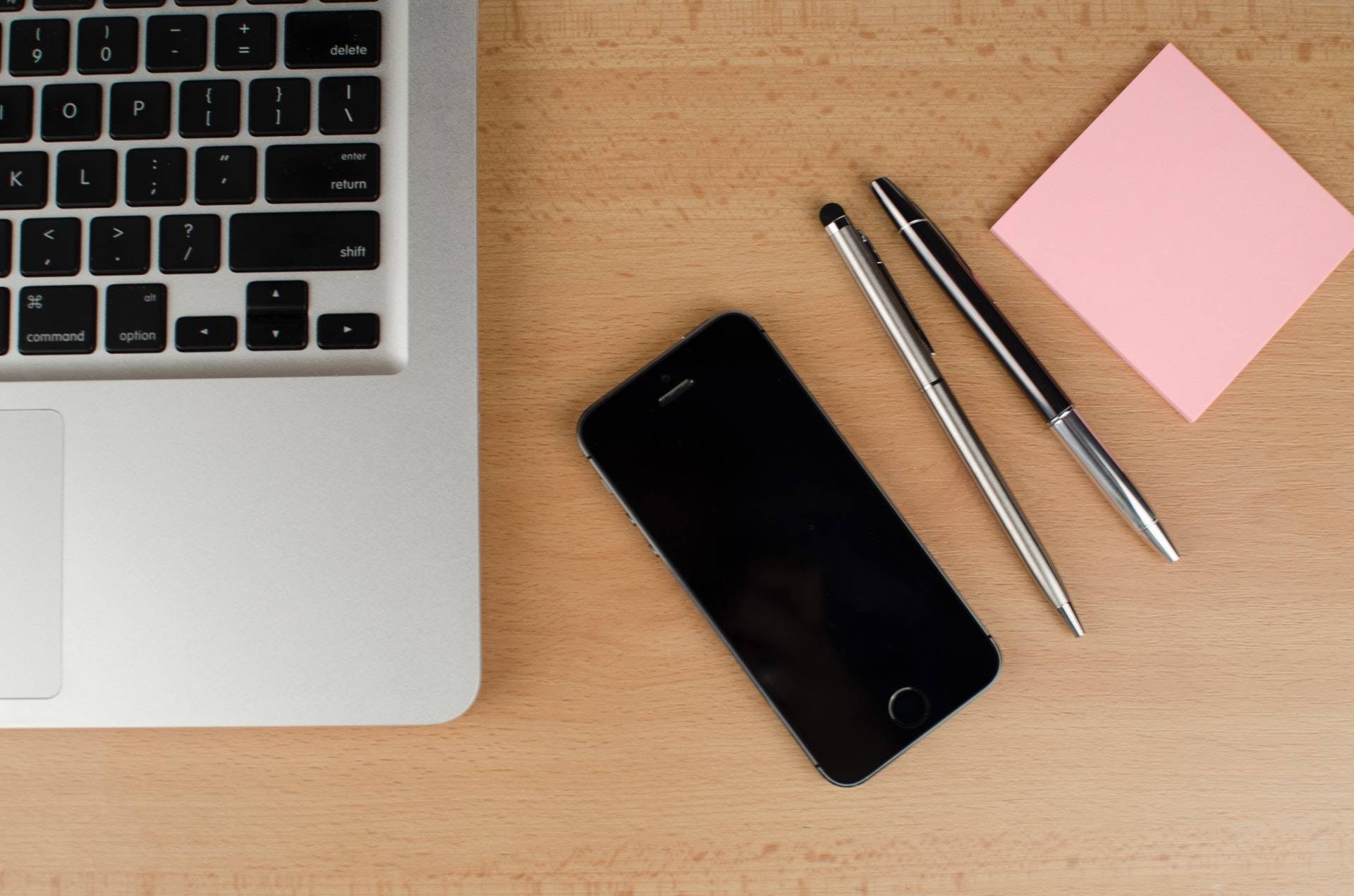 A photograph of a desk with the keyboard of a laptop visible, a phone with a blank screen, two pens, and a pink sticky note pad