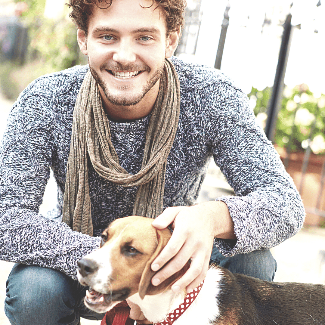 A person wearing a grey knit sweater and brown scarf smiling at the camera. The person is petting a small tri-color beagle that is looking off to the left